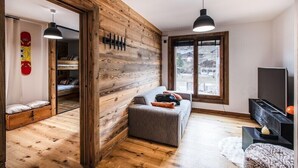A cozy room with a wooden wall, a gray sofa, a TV stand, and a window with a view. - PEARL 201 | LES GETS | 14 pers.  | Central (Les Gets)