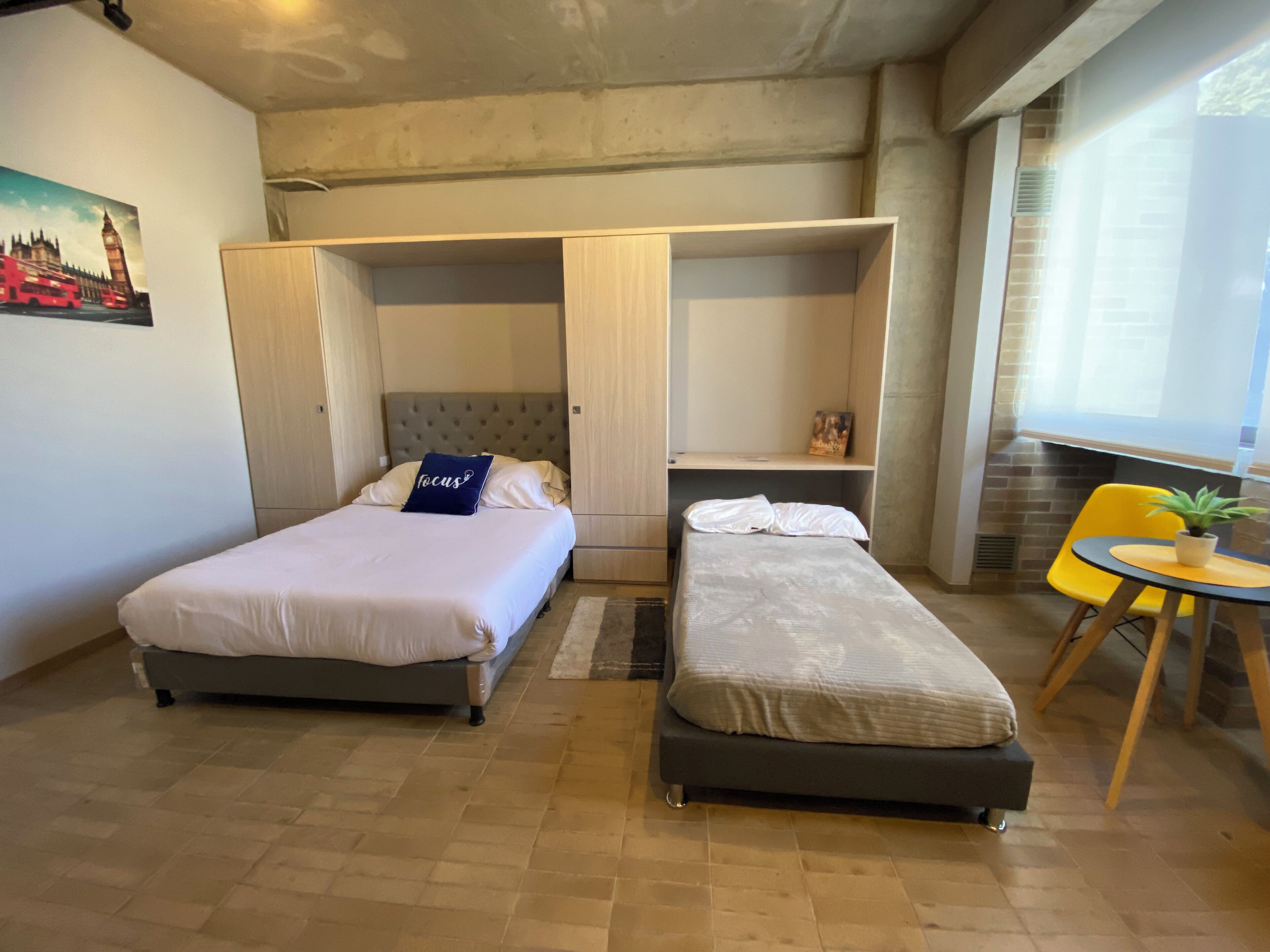 A room with two beds, a small table with a plant, and a window with a view of the outside.