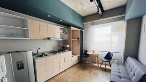 Et kompakt køkken med moderne hårde hvidevarer, et spiseområde med et lille bord og stole, samt en grå sofa.