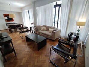 A living room with a sofa, coffee table, and a treadmill. There is a dining table with chairs and a lamp.