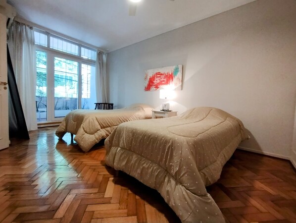 A room with two beds, a window with curtains, a nightstand with a lamp, and a painting on the wall.
