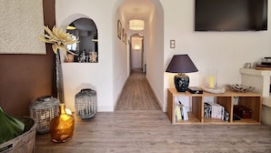 Un pasillo con piso de madera, un televisor montado en la pared, una lámpara sobre una mesita auxiliar y una estantería con libros y objetos decorativos.