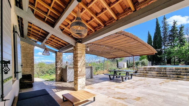 Un patio al aire libre con suelo de piedra, un banco de madera, una mesa de picnic y una pérgola con una lámpara colgante.