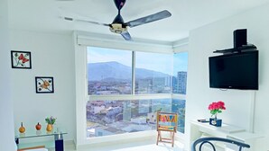 Une pièce lumineuse avec une grande fenêtre, un ventilateur de plafond, un téléviseur à écran plat et une vue sur le paysage urbain.
