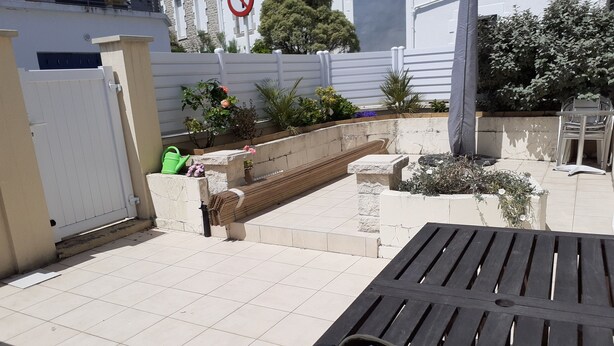 Een patio met betegelde vloer, een houten bank en een tafel met tas en pet.