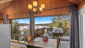 A wooden interior with a dining table, chairs, a chandelier, and a view of a snowy landscape through large windows. - Le P`tit Bonheur - Lac et Nature (Sainte-Lucie-des-Laurentides)