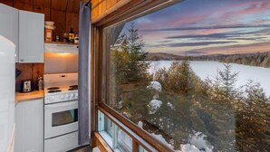 A cabin interior with a kitchen, a large window with a snowy landscape view, and wooden walls. - Le P`tit Bonheur - Lac et Nature (Sainte-Lucie-des-Laurentides)