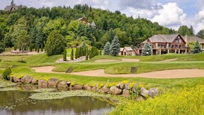 A golf course with a pond, a lodge with a balcony, and a forested hill in the background. - Le P`tit Bonheur - Lac et Nature (Sainte-Lucie-des-Laurentides)