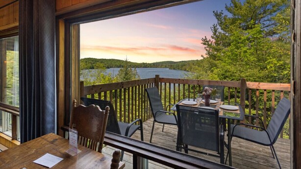 A wooden deck with a table and chairs, overlooking a lake and forested area. - Le P`tit Bonheur - Lac et Nature (Sainte-Lucie-des-Laurentides)