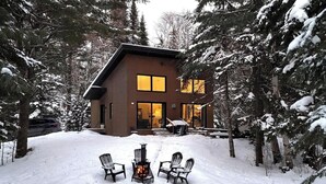 Una cabaña con chimenea, terreno cubierto de nieve y árboles circundantes.