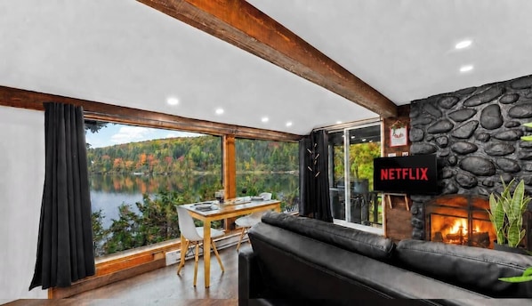 Une pièce chaleureuse avec une cheminée en pierre, un logo Netflix, une grande fenêtre donnant sur un lac et un canapé en cuir noir.