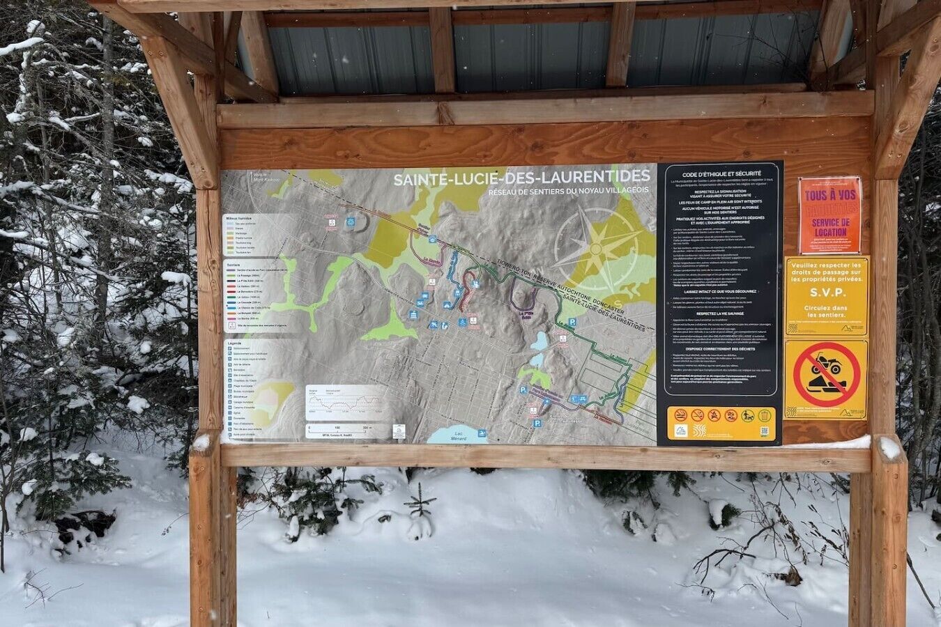 Ein hölzernes Infobrett mit einer Karte und Text, umgeben von schneebedeckten Bäumen.