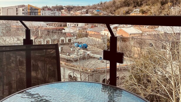 A balcony with a glass table and a view of a town with buildings and trees. - City Heart Luxury Apartment  (Kutaisi)