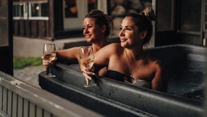 Dos personas en un jacuzzi con un muro de piedra y techo de madera al fondo.