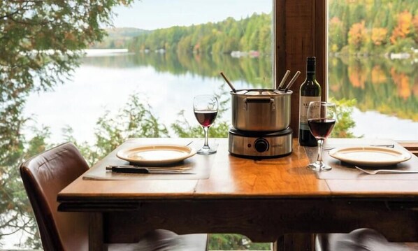 Ein Tisch ist mit Tellern, Weingläsern, einem Topf und einer Flasche gedeckt, mit Blick auf einen See und Wald.