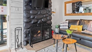 A living room with a stone fireplace, a sofa with cushions, a television mounted on the wall, and a small table with a plant.