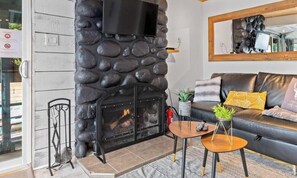 Un salon avec une cheminée en pierre, un canapé avec des coussins, un téléviseur fixé au mur et une petite table sur laquelle est posée une plante.