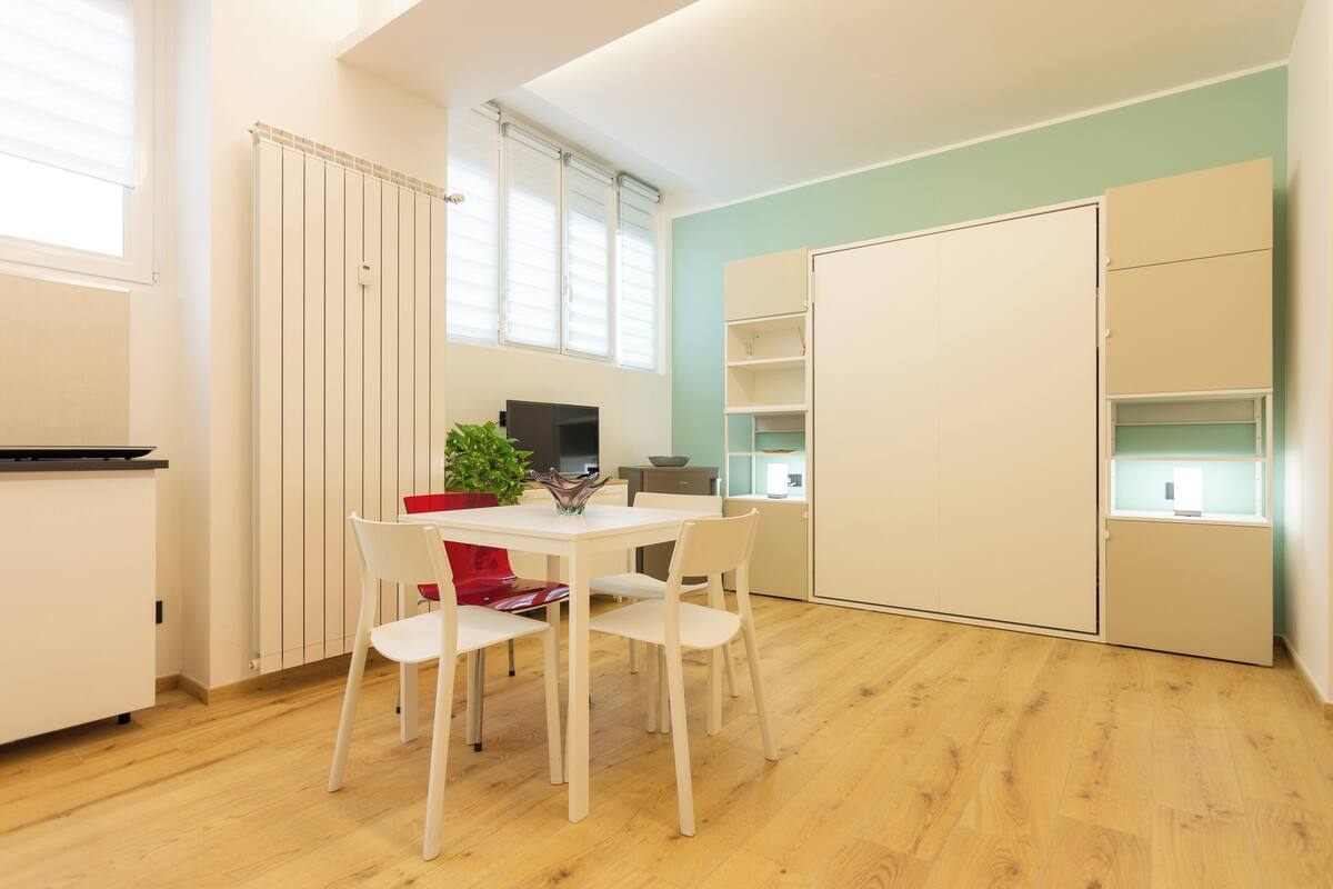 A modern kitchen with a dining table, chairs, and a red chair. There is a plant on the table and a TV on the wall.