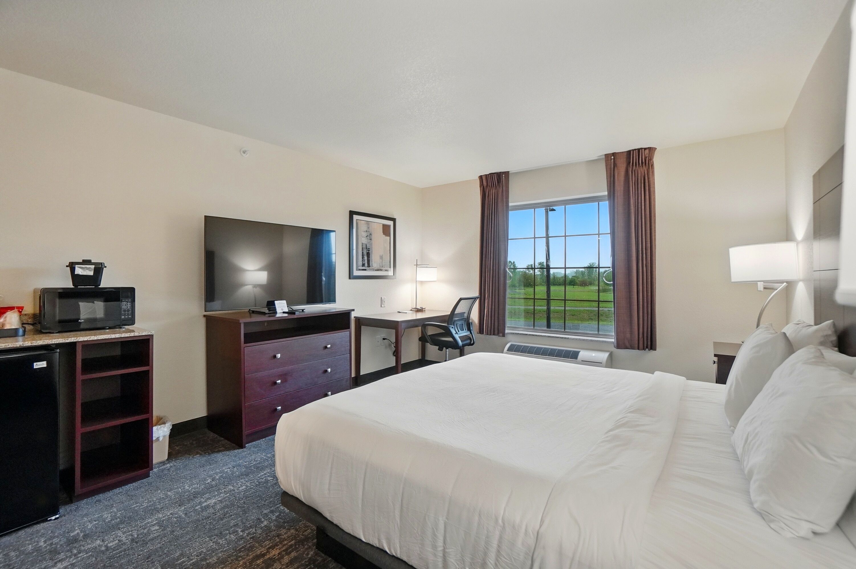 A hotel room with a large bed, a desk with a chair, a television, a microwave, and a window with curtains.
