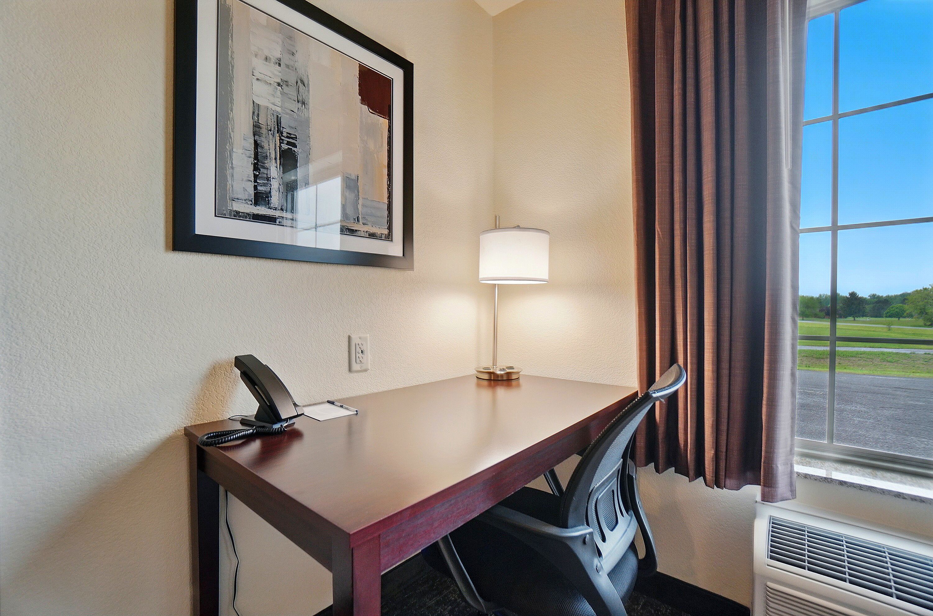Habitación de hotel con escritorio, una obra de arte abstracta enmarcada, una lámpara y vista a un estacionamiento a través de una ventana.