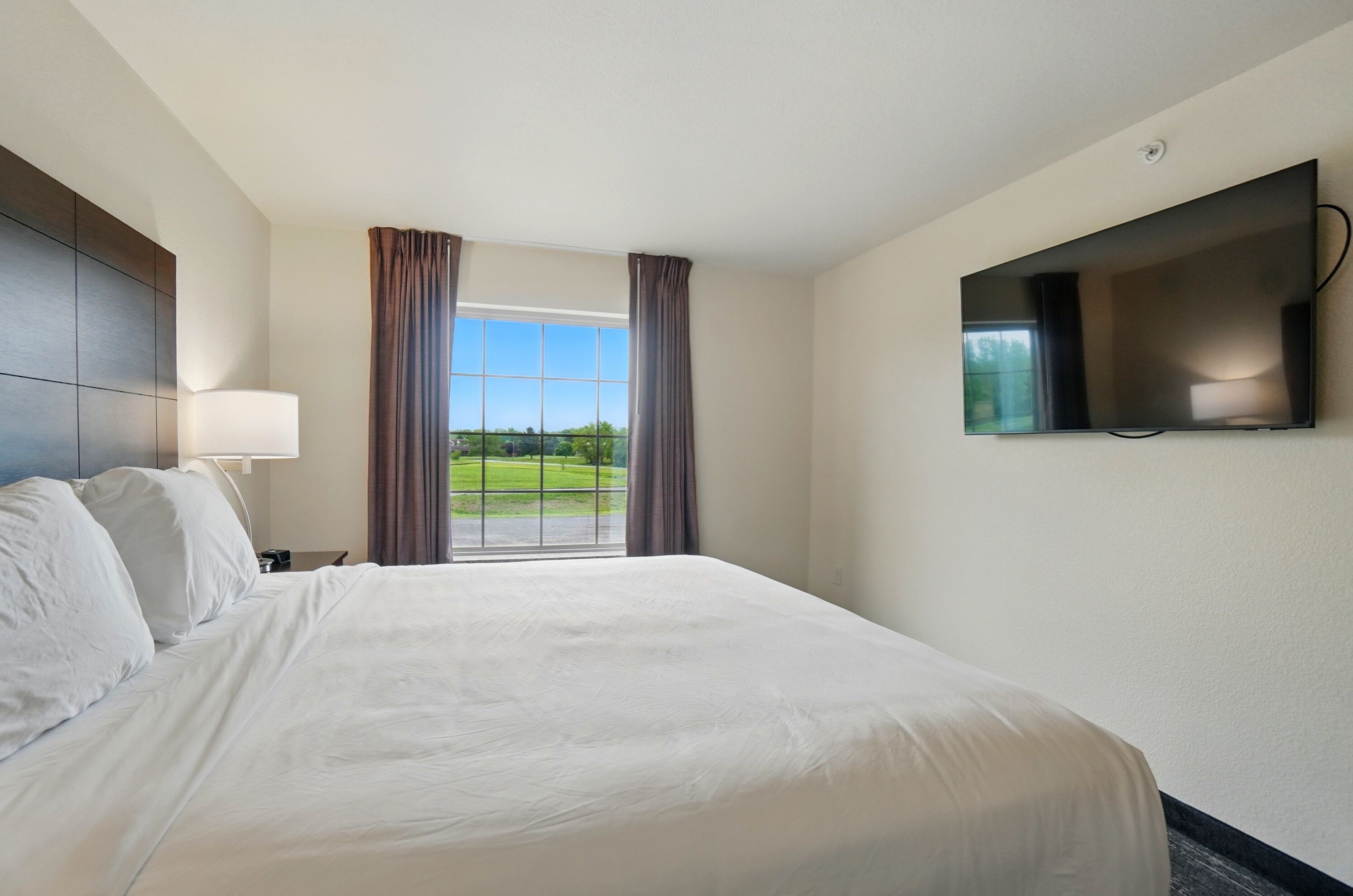 A hotel room with a large bed, a flat-screen TV mounted on the wall, and a window with curtains.