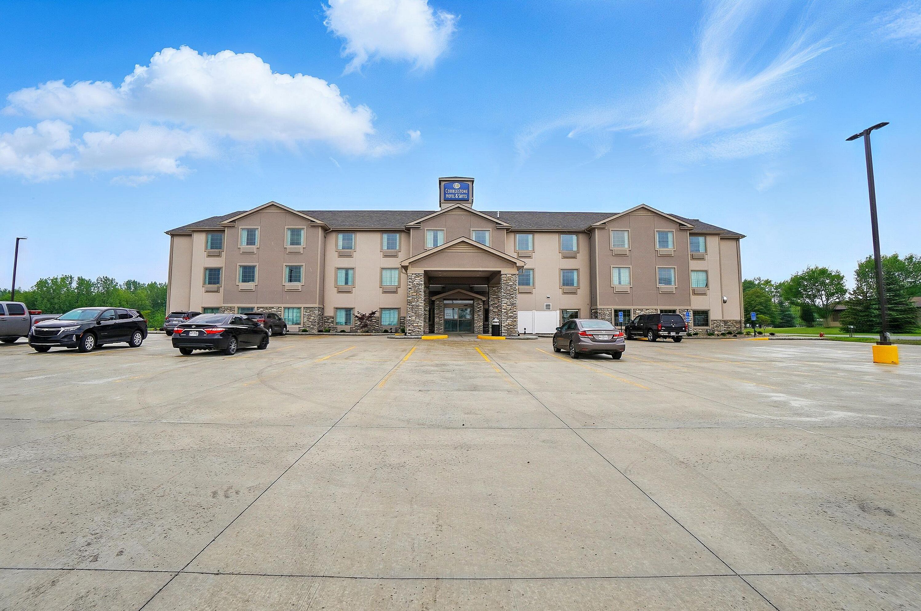 Un hotel de varios pisos con entrada principal, estacionamiento y un cielo azul despejado.