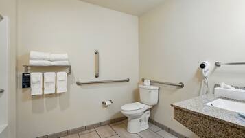 Une salle de bain équipée de toilettes, de barres d’appui, d’un porte-serviettes, d’un lavabo et d’un sèche-cheveux.