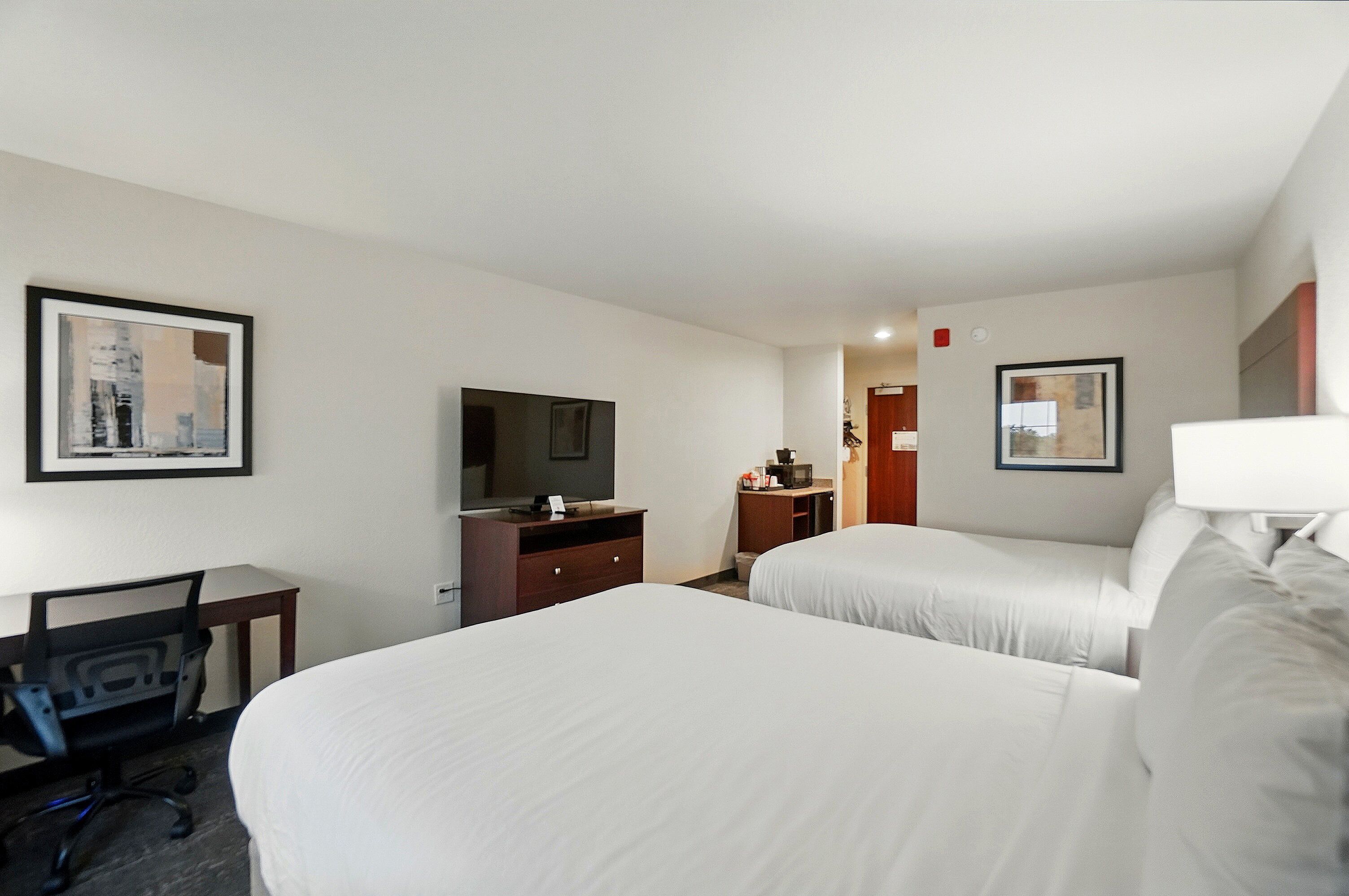 A hotel room with two beds, a desk with a chair, a television, and a small kitchenette.