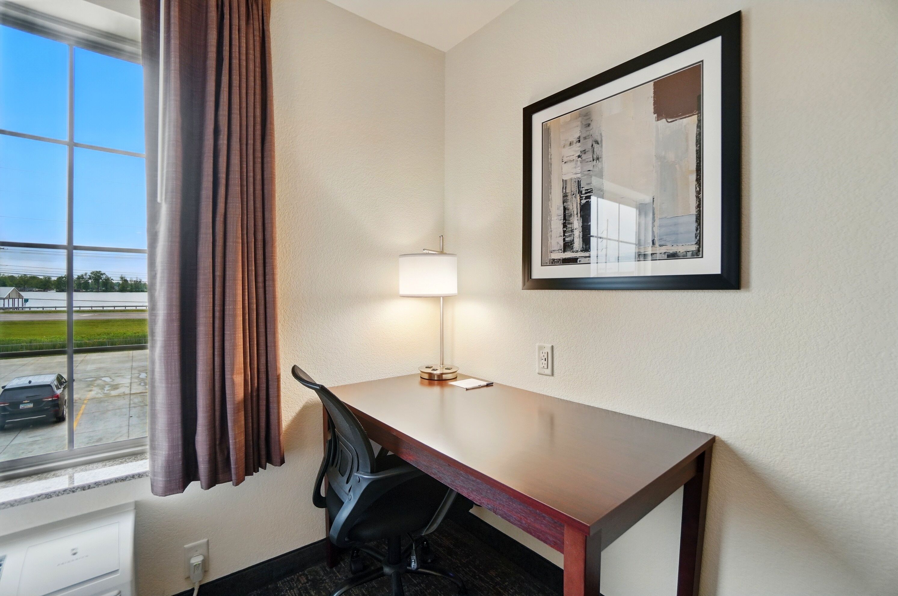 A hotel room with a desk, chair, lamp, and a framed abstract painting on the wall.