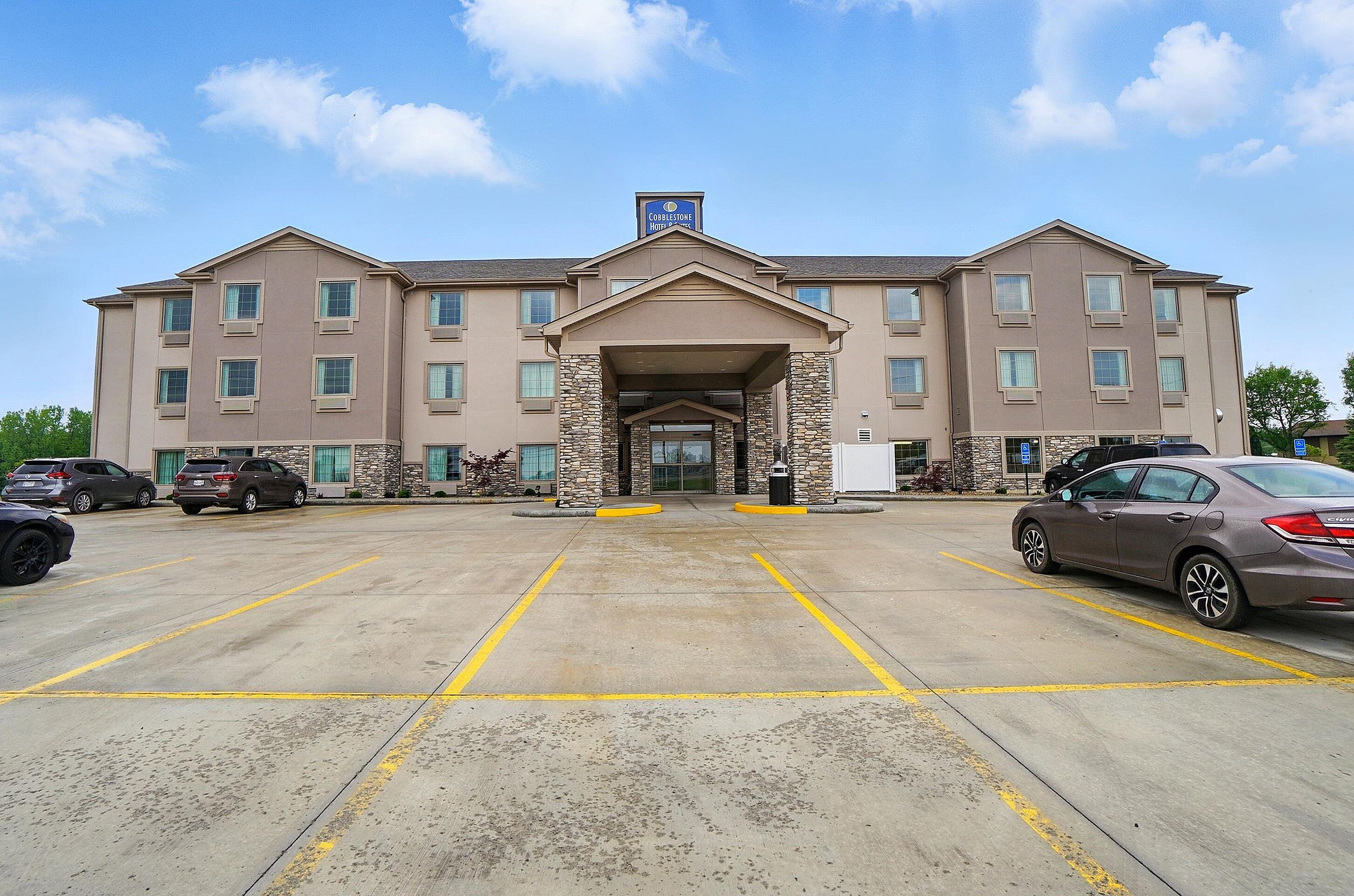 Un hotel con entrada central, varios espacios de estacionamiento y cielo despejado.