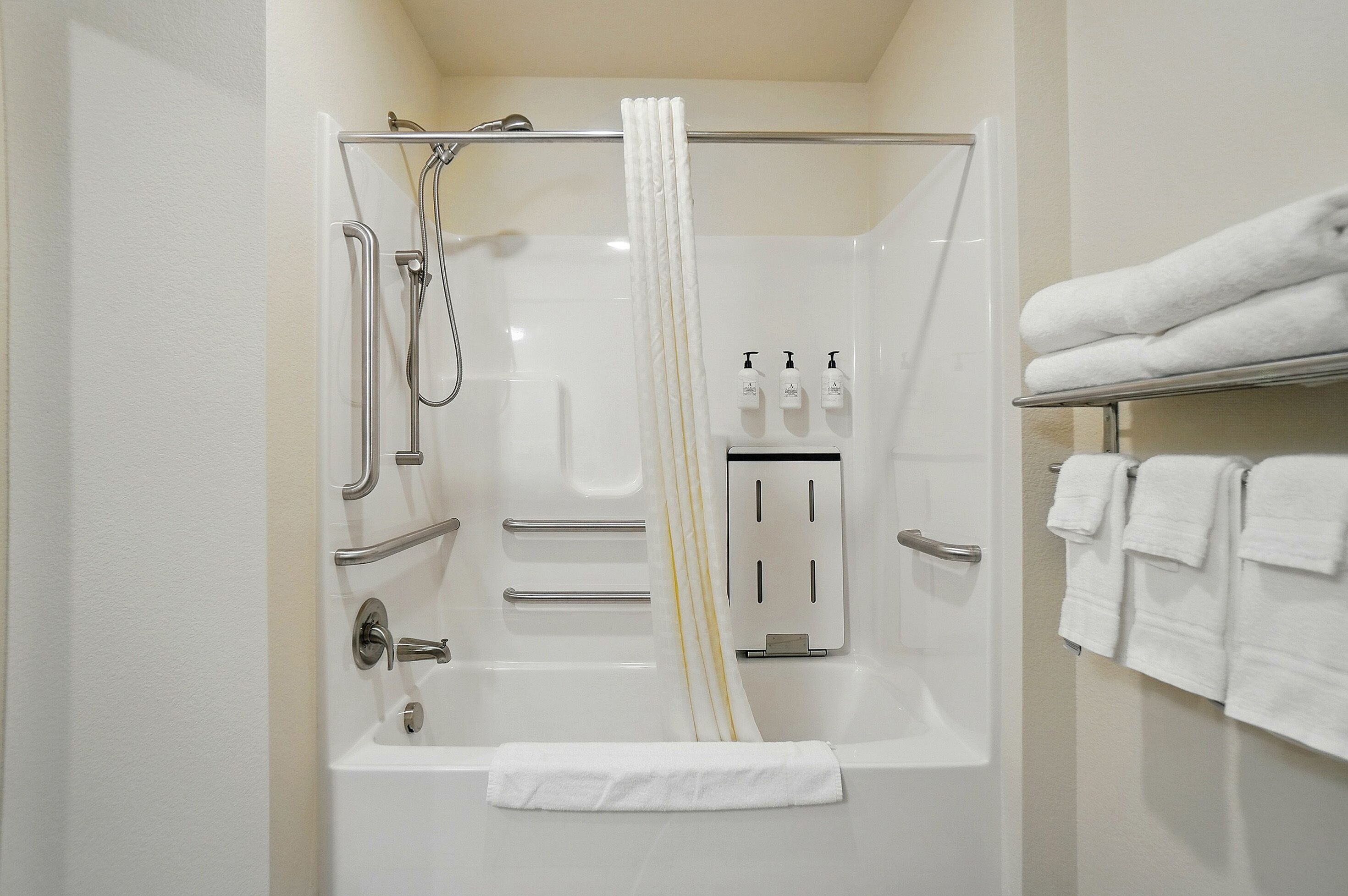 Un baño moderno con bañera, ducha y barras de apoyo. Hay toallas en un perchero y dispensadores de jabón en la pared.