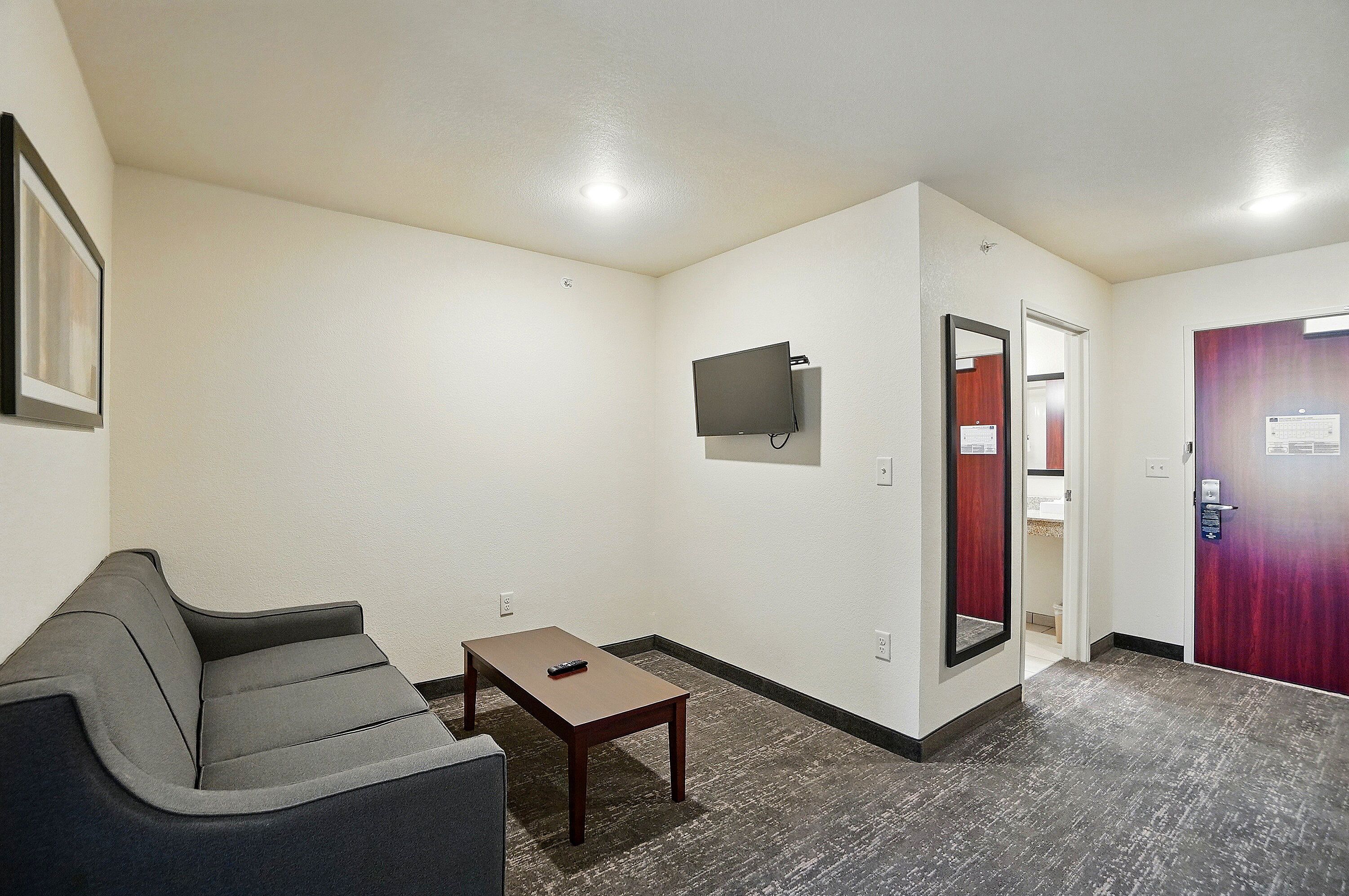 A hotel room with a gray sofa, a small wooden table, a flat-screen TV mounted on the wall, and a door with a red frame.
