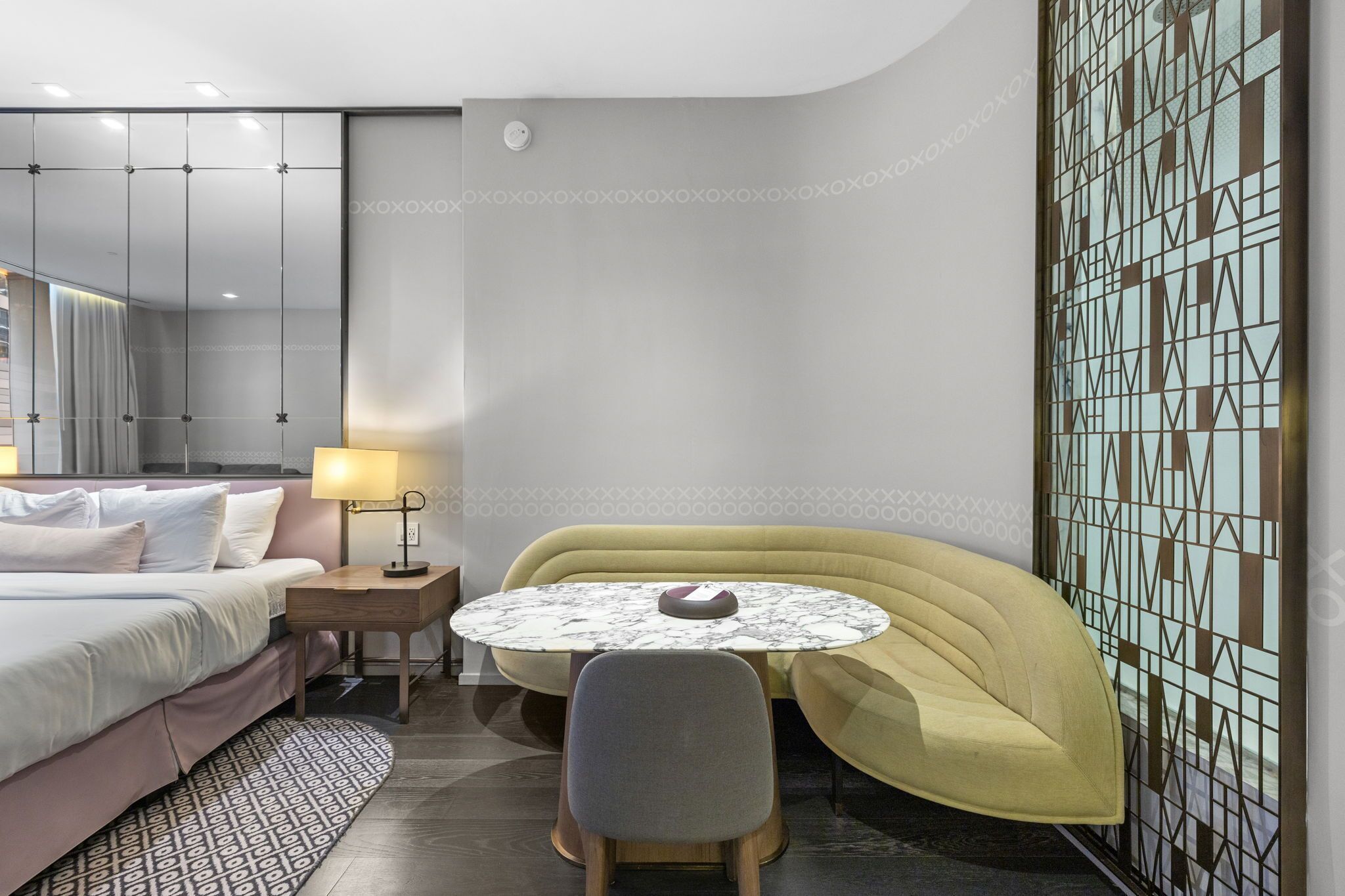 Une chambre d’hôtel moderne comprenant un lit, une table ronde avec une chaise, une petite table de chevet avec une lampe et un panneau décoratif mural.