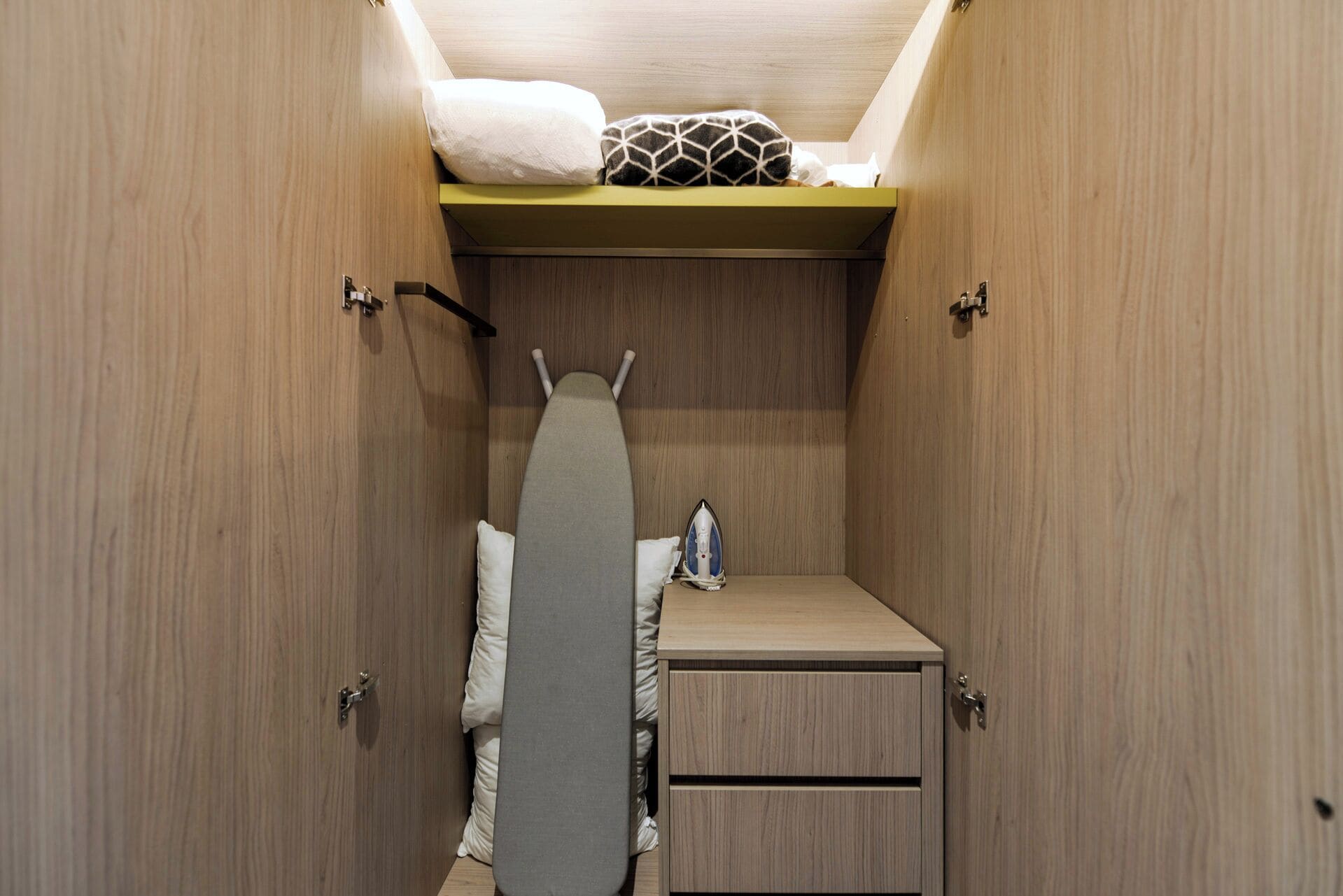 A compact bedroom with a bed, a small dresser, a shelf with pillows, and an ironing board.