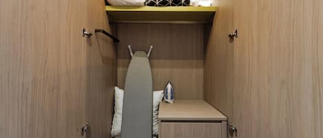 A compact bedroom with a bed, a small dresser, a shelf with pillows, and an ironing board.