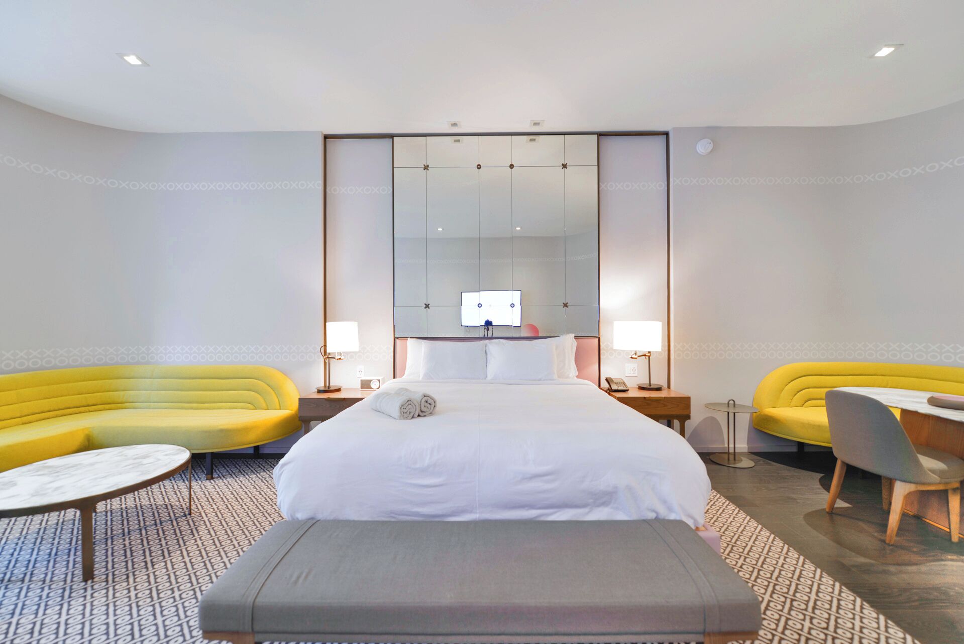 A modern hotel room with a large bed, two yellow sofas, a small round table, and a grey bench.
