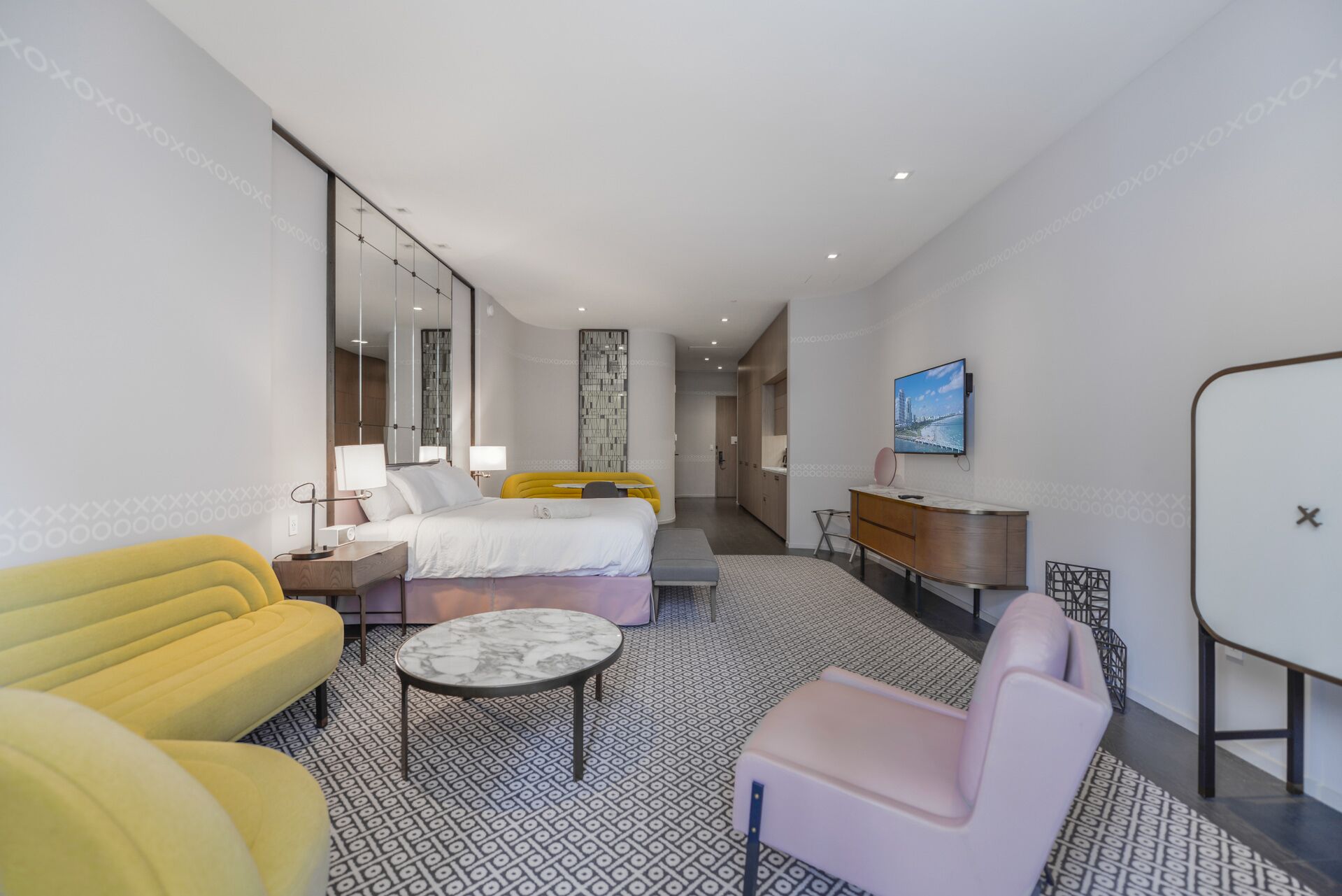 A modern hotel room with a bed, a desk, a TV, and a seating area with a patterned floor.