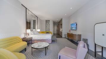 A modern hotel room with a bed, a desk, a TV, and a seating area with a patterned floor.