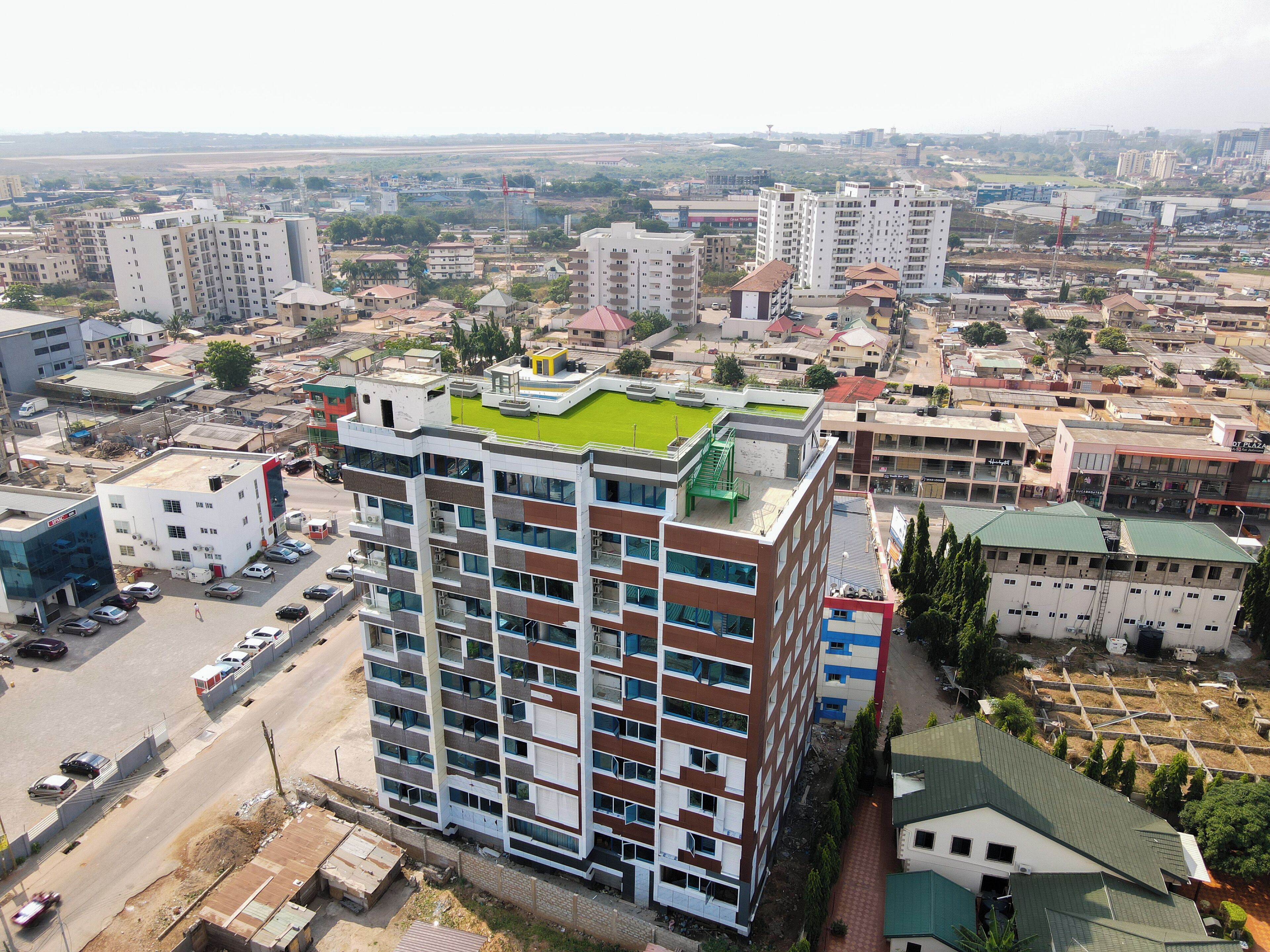 Un edificio de varios pisos con una terraza verde en la azotea, rodeado de otros edificios y un estacionamiento.