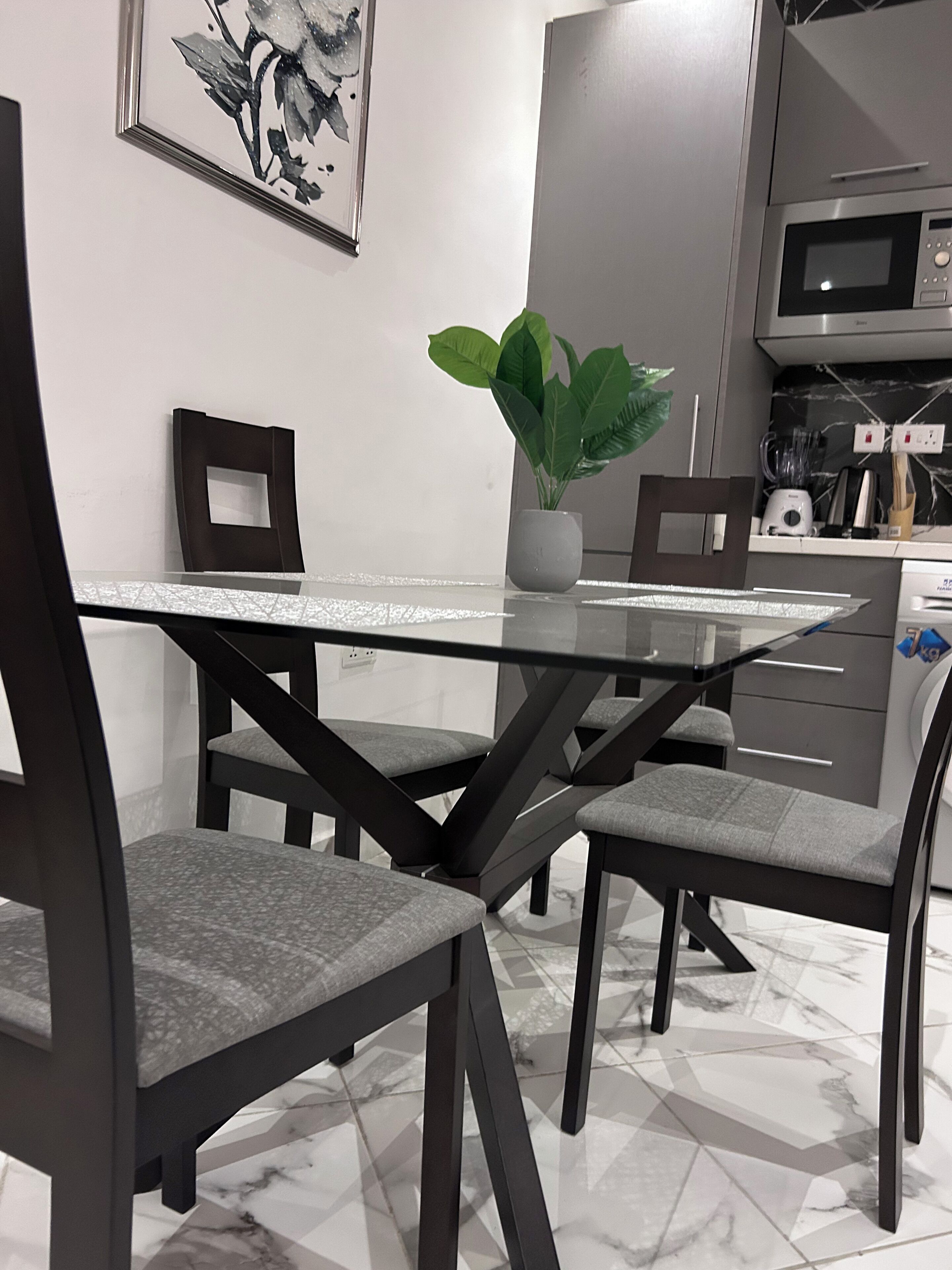 A modern dining area with a glass table and dark chairs, a potted plant, and a kitchen in the background.