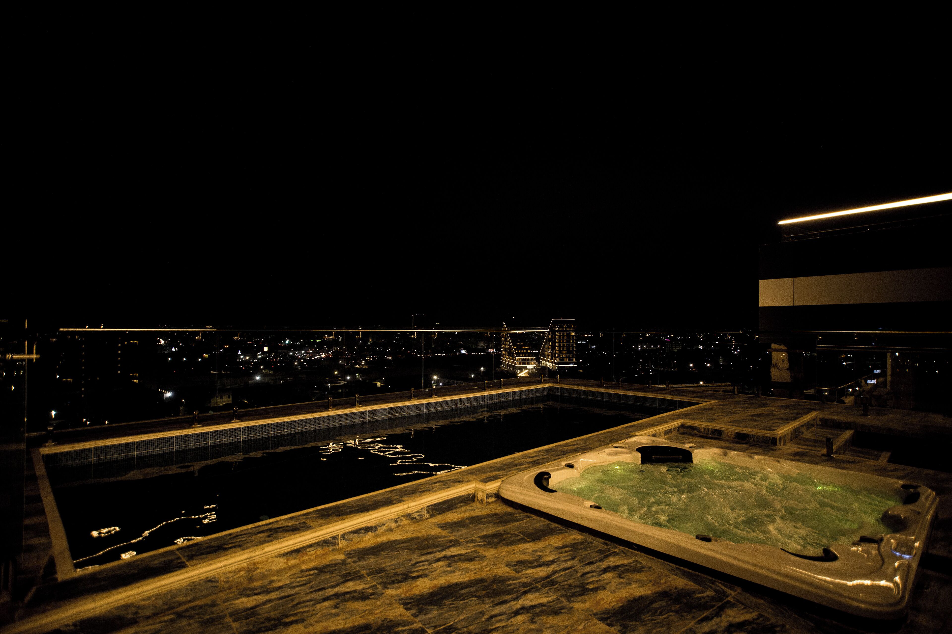 A rooftop pool area with a hot tub and a view of the city at night.