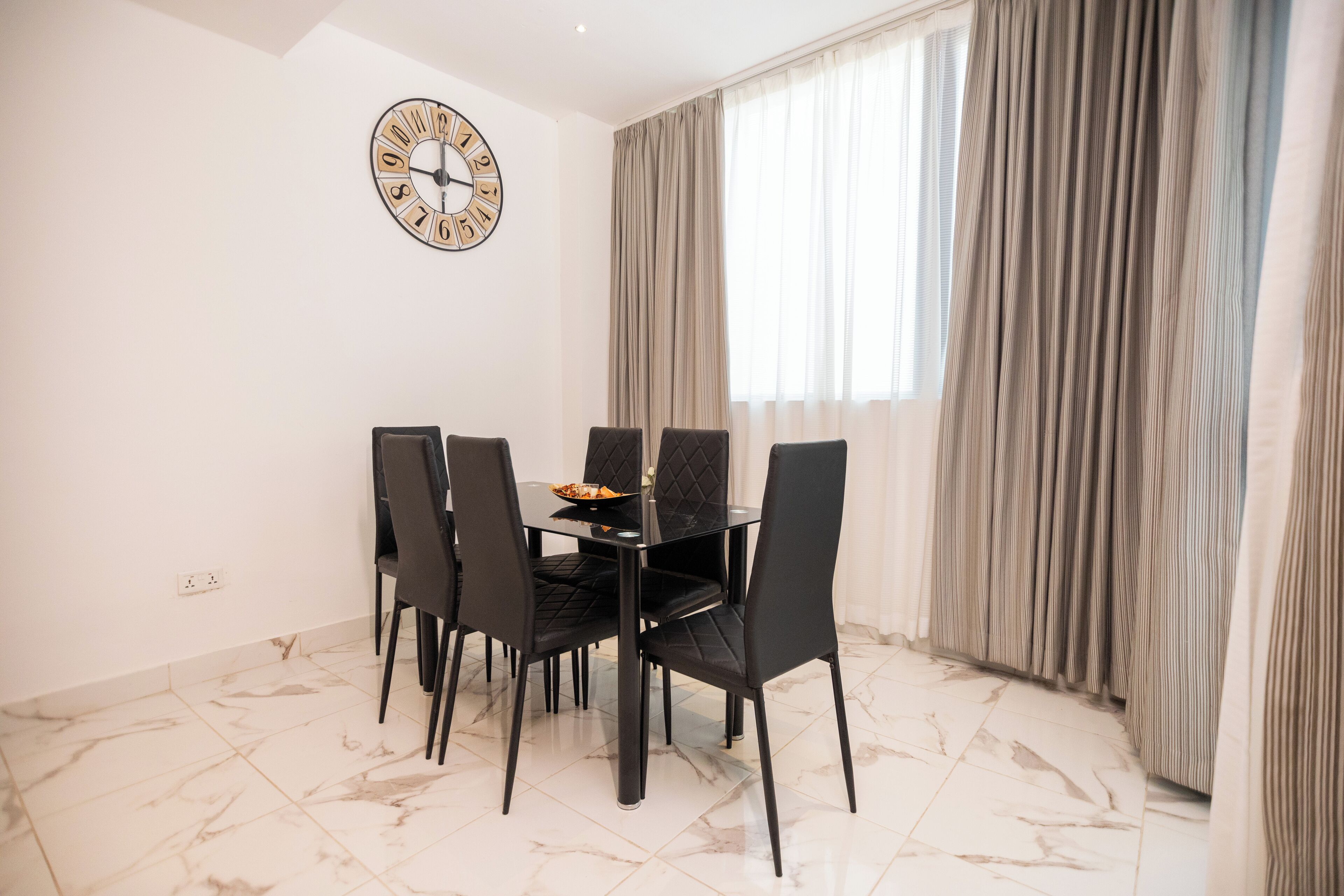 Uma área de jantar com uma mesa redonda, cadeiras pretas e um relógio na parede.