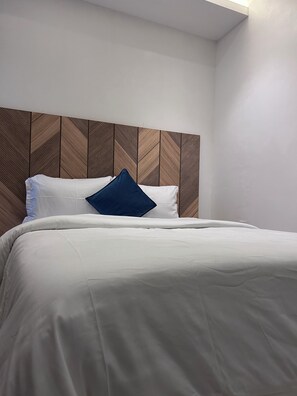 A neatly made bed with white bedding and a blue pillow, against a wall with a wooden panel design. - BOT PROPERTIES (Accra)