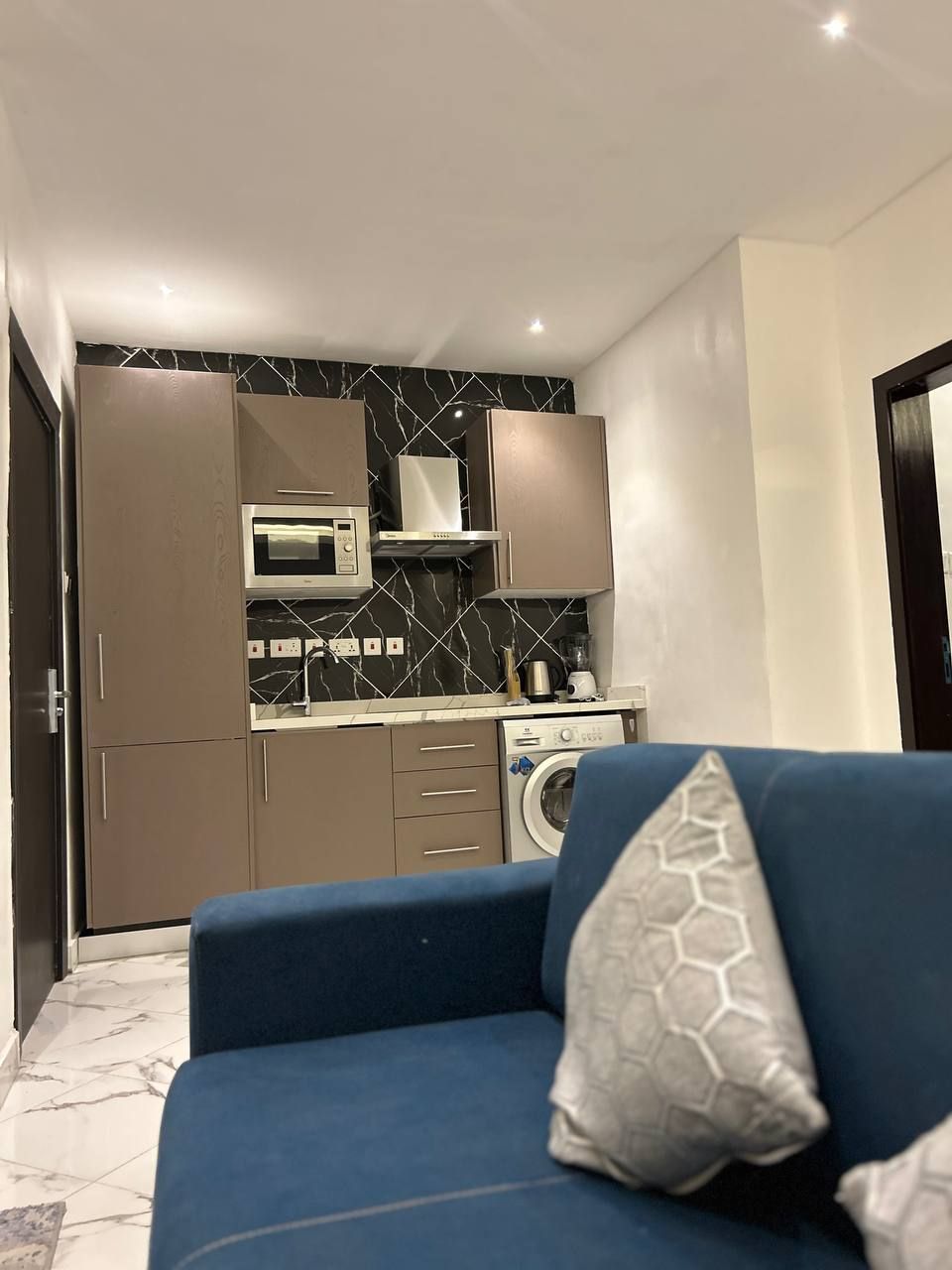 A compact living space with a kitchenette, a washing machine, and a blue sofa with patterned cushions.