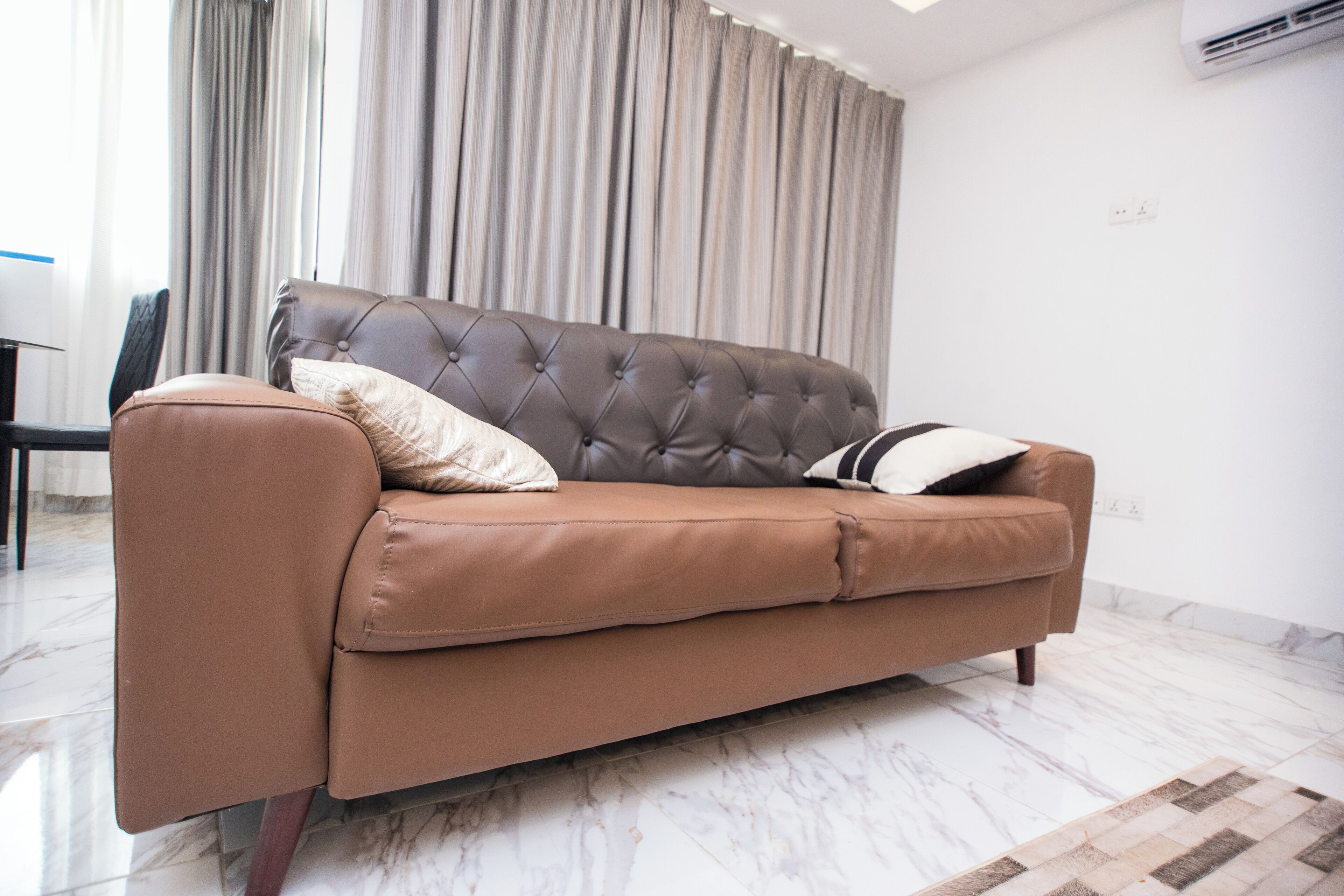 A brown leather sofa with a tufted backrest and two pillows, positioned in a room with a marble floor and a large window with curtains.
