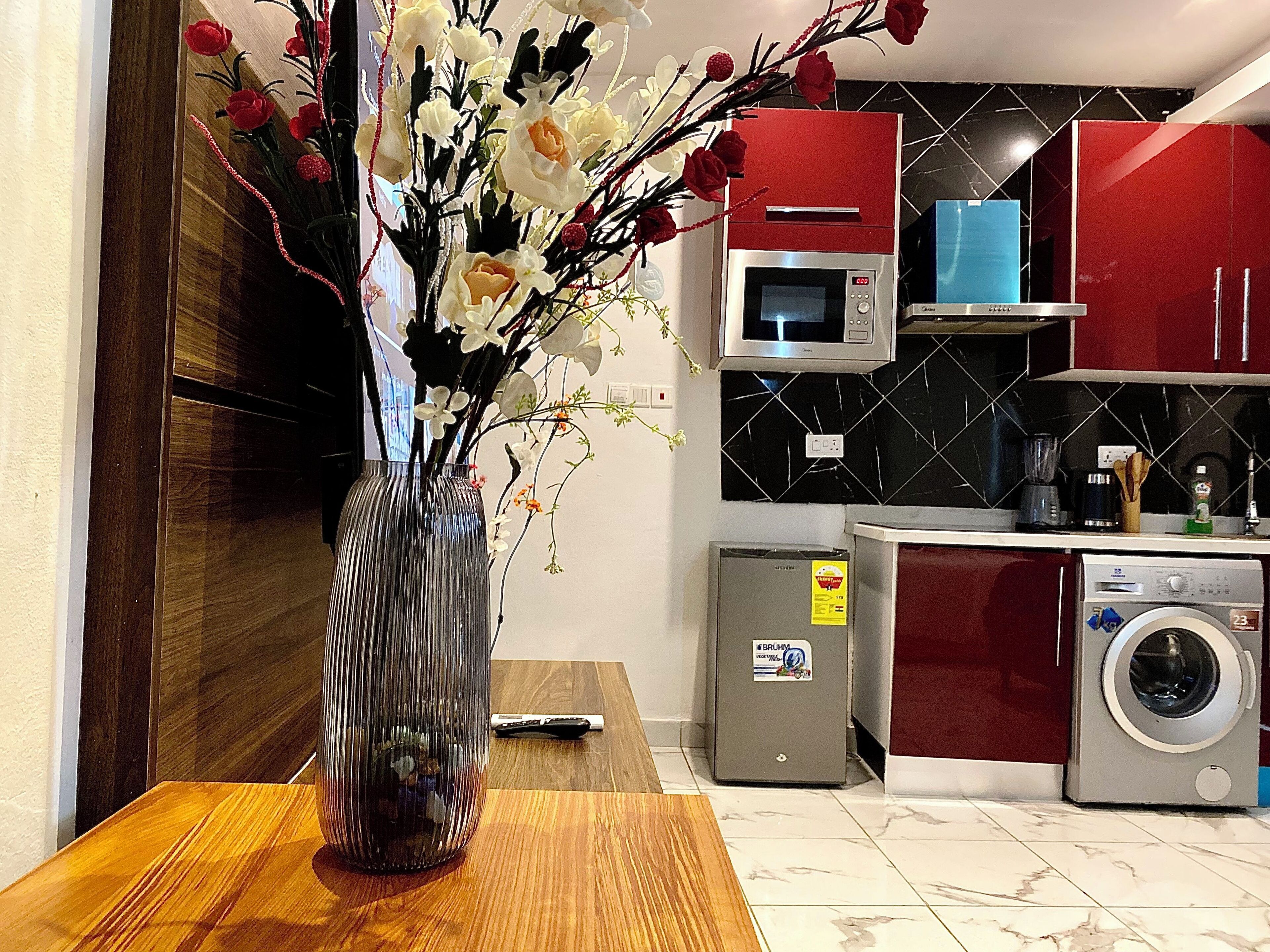 A modern kitchen with a wooden table, a vase with flowers, a microwave, a washing machine, and red cabinets.