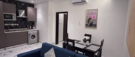 A modern living room with a blue sofa, a dining table with chairs, a kitchen area with a microwave and washing machine, and an air conditioning unit on the wall.