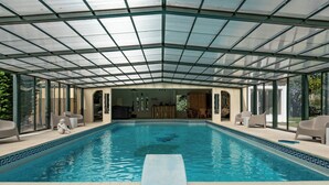 Une grande piscine intérieure dotée d’un toit en verre, entourée de chaises longues et d’une terrasse en bois.