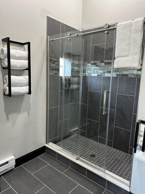 A modern shower area with a glass door, dark tiles, and a towel rack with folded towels. - Big Cat Bungalows at The Haven (Chittenango)