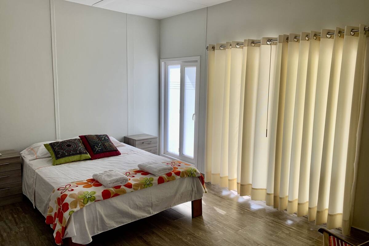 A bedroom with a bed, a nightstand, a window with curtains, and a wooden floor.